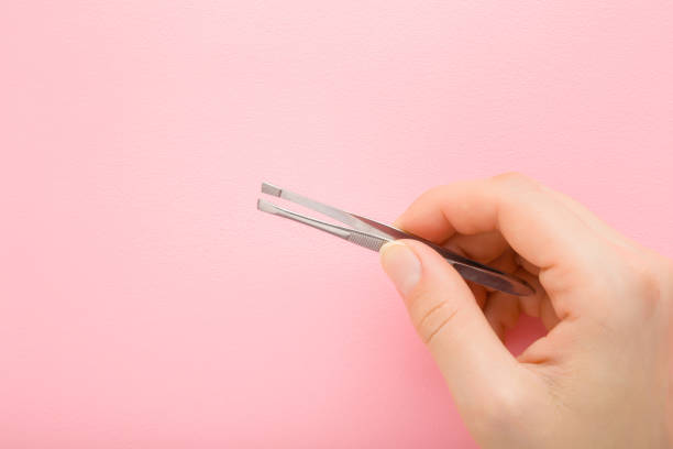 Young adult woman fingers holding tweezers on light pink background. Pastel color. Closeup. Top down view. stock photo