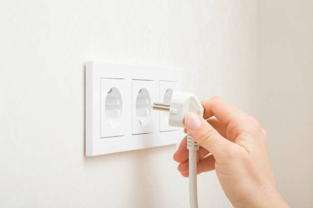 Young adult woman hand holding and plugging white electrical plug in wall outlet socket at home. Closeup. Side view. stock photo