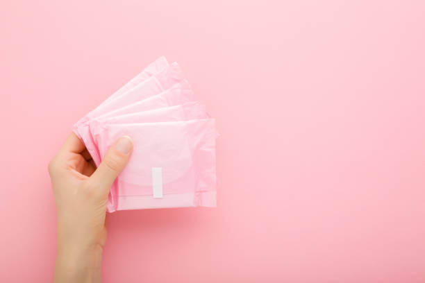 Young adult woman hand holding packs of sanitary towel on light pink table background. Pastel color. Closeup. Empty place for text. Top down view. stock photo