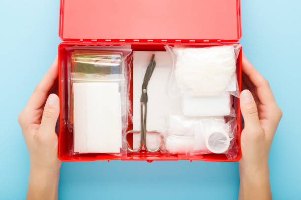 Young adult woman hands holding opened red first aid kit box with different medical accessories in packages on light blue table background. Pastel color. Closeup. Point of view shot. Top down view. stock photo