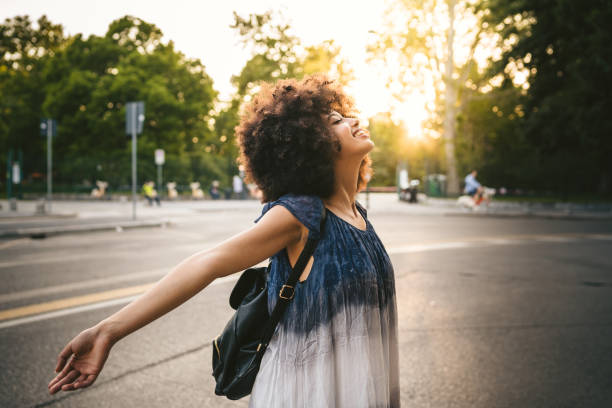 Young adult woman relaxing on the street at sunset in the city in summer opening your arms and looking upwards with closed eyes and smiling - Millennial is free and carefree stock photo