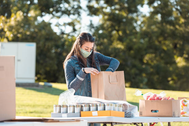 joven mujer adulta usa máscara mientras clasifica los alimentos donados - giving tuesday fotografías e imágenes de stock