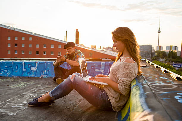 young adults sitting on a roof, back lit young woman sitting on a roof in Berlin and works at her laptop, in the background the skyline from Berlin with the television tower central berlin stock pictures, royalty-free photos & images