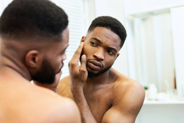 jeune type américain africain contrôlant sa peau de visage dans le miroir - homme miroir photos et images de collection