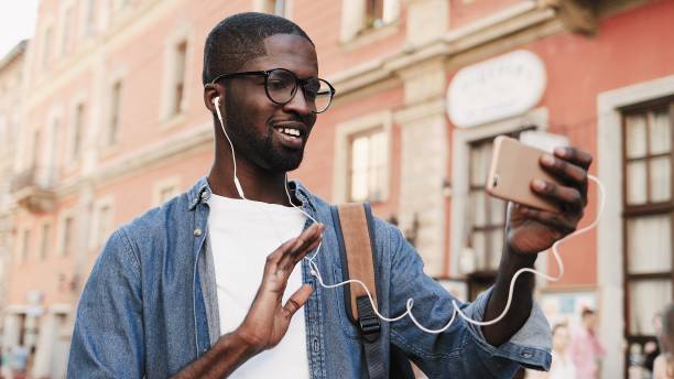 Young African American man is talking on video call from his phone on street using headphones. Guy waves his hand goodbye on video call. stock photo