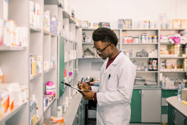 jeune pharmacien d'homme d'afro-américain restant à l'intérieur de la pharmacie. spécialiste d'homme de la pharmacie prenant des notes sur le presse-papiers pendant l'inventaire - pharmacien photos et images de collection