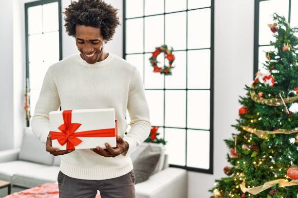 young african american man unboxing gift standing by christmas tree at home. - unbox stockfoto's en -beelden