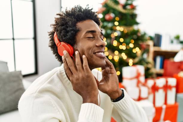 Young african american man using headphones sitting by christmas tree at home. stock photo