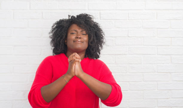 Young african american plus size woman over white brick wall begging and praying with hands together with hope expression on face very emotional and worried. Asking for forgiveness. Religion concept.  prayer request stock pictures, royalty-free photos & images