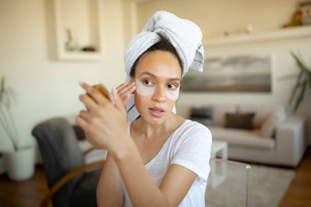 Young African American woman applying makeup on her face Attractive young woman of African American ethnicity, holding a small vanity mirror and taking care of herself, having her own spa treatment at home applying face cream stock pictures, royalty-free photos & images