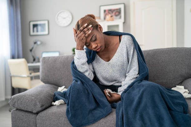Young african american woman, sitting on the sofa at home, having a fever  and  touching her forehead to check her temperature stock photo