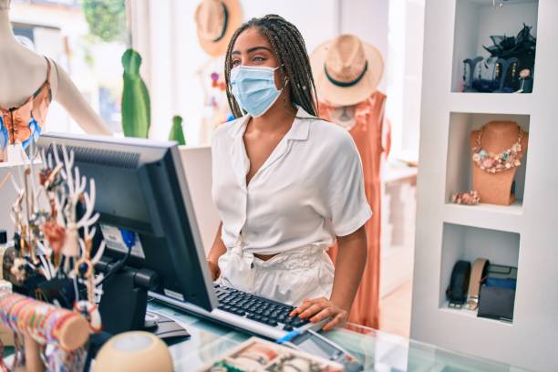 Young african american woman smiling happy working at the till wearing coronavirus safety mask at retail shop Young african american woman smiling happy working at the till wearing coronavirus safety mask at retail shop boutique stock pictures, royalty-free photos & images