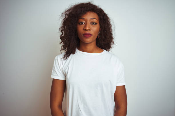 jonge afrikaanse amerikaanse vrouw die t-shirt draagt dat zich over geïsoleerde witte achtergrond bevindt ontspannen met ernstige uitdrukking op gezicht. eenvoudig en natuurlijk kijken naar de camera. - afro amerikaanse etniciteit stockfoto's en -beelden