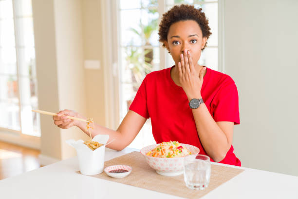 Young african american woman with afro hair eating asian food at home cover mouth with hand shocked with shame for mistake, expression of fear, scared in silence, secret concept Young african american woman with afro hair eating asian food at home cover mouth with hand shocked with shame for mistake, expression of fear, scared in silence, secret concept how do you say shut up in japanese stock pictures, royalty-free photos & images
