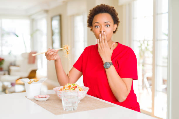 Young african american woman with afro hair eating asian food at home cover mouth with hand shocked with shame for mistake, expression of fear, scared in silence, secret concept Young african american woman with afro hair eating asian food at home cover mouth with hand shocked with shame for mistake, expression of fear, scared in silence, secret concept how do you say shut up in japanese stock pictures, royalty-free photos & images