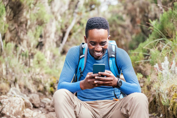 Young African ethnicity man video chatting via cellphone with somebody and cheerfully laughing as he having a hiking walk in the forest. Happy people, network technology, or traveling concept stock photo