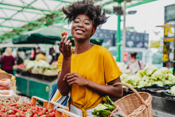 Young African woman buying fruits at the market A happy smiling African-American woman is holding a basket full of groceries farmer's market stock pictures, royalty-free photos & images