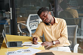 istock Young African-American businessman sitting at desk in office and using mobilephone while discussing details of campaign 1335084864