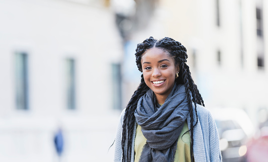 A young African-American woman with long braided hair, walking in the city, smiling at the camera.