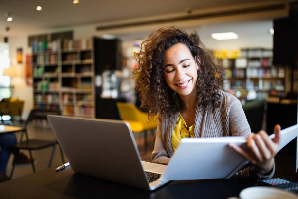 jonge afro amerikaanse vrouwenzitting bij lijst met boeken en laptop voor het vinden van informatie - studerende stockfoto's en -beelden
