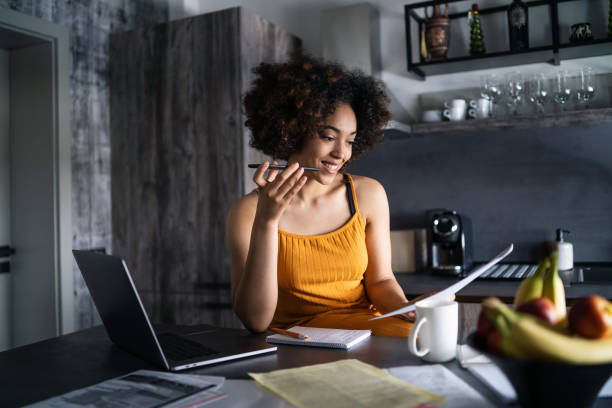 Young afro woman having a work day at home office, sitting in the kitchen, checking documents and talking on the phone stock photo