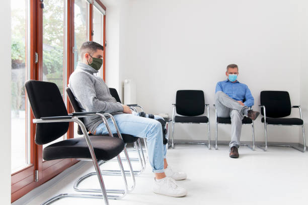 junger und reifer mann mit gesichtsmasken sitzt in einem wartezimmer eines krankenhauses oder büros - abwarten stock-fotos und bilder