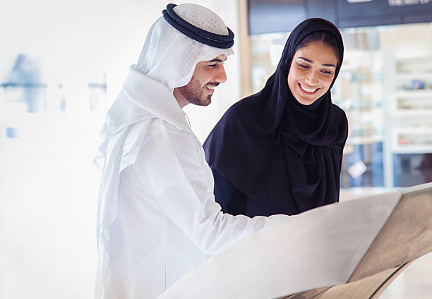 Young Arab couple using information display at mall Emirati couple with traditional clothes using electronic info display in a shopping mall. abaya clothing stock pictures, royalty-free photos & images