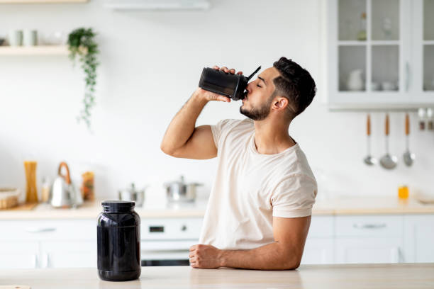Young Arab guy drinking protein shake from bottle at kitchen, copy space. Body care concept stock photo