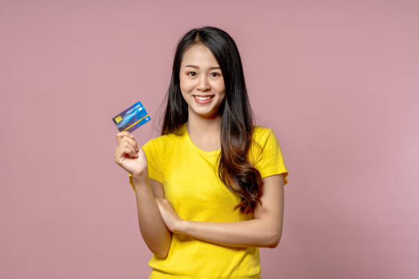 年輕的亞洲美女在黃色襯衫微笑，並顯示信用卡在手感覺信任和信心支付與工作室拍攝的粉紅色背景。 - show up kamerki 個照片及圖片檔