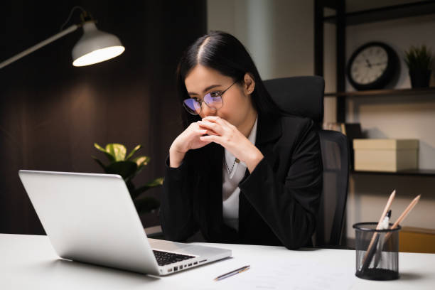 Young asian business woman working at late night. She was very busy checking paperwork and had to send the work before the deadline, feeling stressed. Sitting in the dark office at night. stock photo