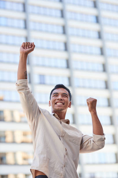A young Asian businessman celebrates with arms raised in front of a skyscraper stock photo