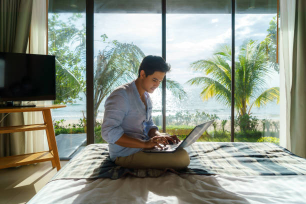 Young Asian businessman sitting in bed and working with laptop in room at resort near sea during a summer vacation holiday travel. stock photo