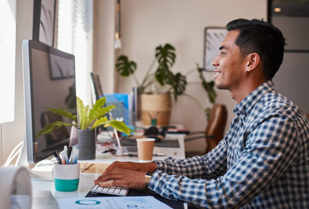 A young Asian businessman types at his desk on the computer while smiling stock photo