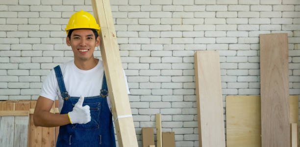 Young asian carpenter wearing a yellow hardhat, carrying wood for making furniture in the workshop room. stock photo