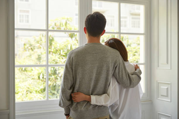 young asian couple staying at home rear view of a young asian couple looking out of window during staying at home order arm around stock pictures, royalty-free photos & images