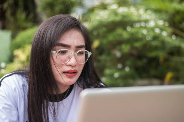 A young asian female with glasses is slightly confused at what she is reading or reviewing on her laptop. Either chatting on social media or studying for an online class. stock photo