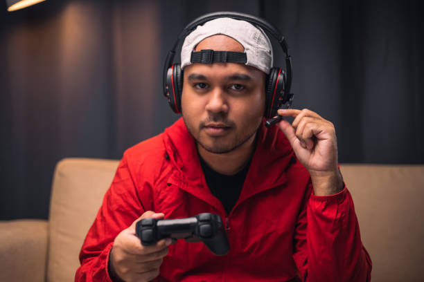 Young asian man in red jacket wearing headset playing game. Indian Gamer holding joystick play video game online at home. stock photo