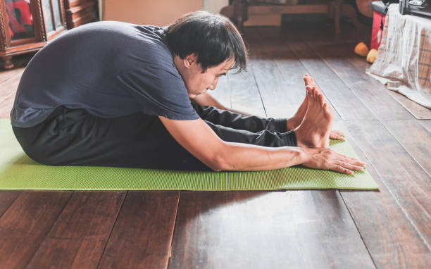 Young Asian man practicing yoga, sitting in Seated forward bend exercise, paschimottanasana pose on green mat yoga at wooden home. Healthy living stock photo