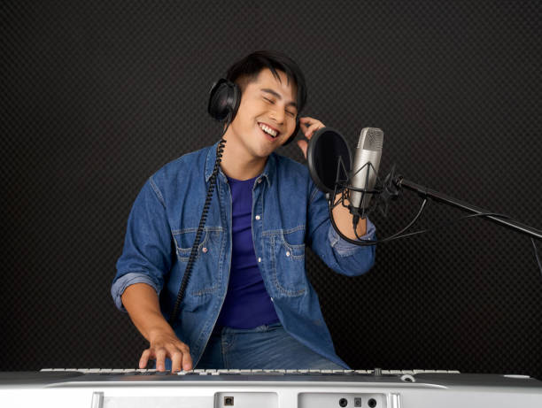 Young asian man with headphones playing an electric keyboard in front of black soundproofing walls. Musicians producing music in professional recording studio. stock photo