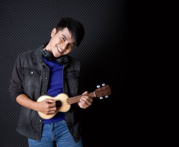 Young asian man with headphones playing an Ukulele guitar in front of black soundproofing walls. Musicians producing music in professional recording studio. stock photo