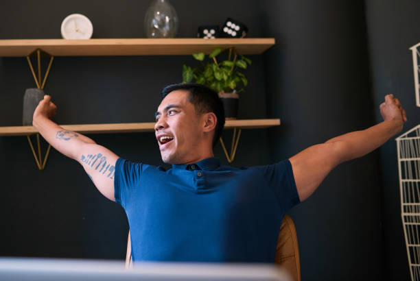 A young Asian man with tattoos stretches at his desk and yawns in the office stock photo