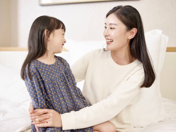 young asian mom enjoying time together with daughter young asian mother and elementary age daughter sitting in bed relaxing playing chatting at home asian mother talking with daughter stock pictures, royalty-free photos & images