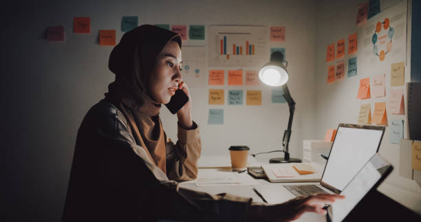 pengusaha muda asia mengenakan jilbab bekerja di laptop dan menggunakan tablet untuk melakukan percakapan telepon sambil duduk di meja di kantor pada malam hari. - mengetik gelap potret stok, foto, & gambar bebas royalti