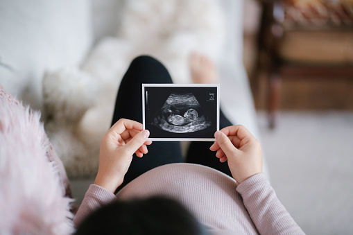 Young Asian pregnant woman lying on sofa at home, looking at the ultrasound scan photo of her baby. Mother-to-be. Expecting a new life concept