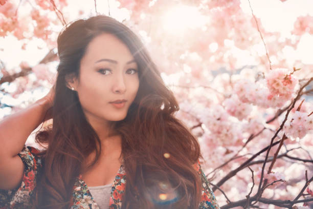 Young Asian woman, in a floral pattern dress, in front of blossoming cherry tree in the spring time stock photo