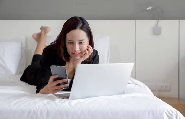 Young asian woman in black suit reading text message on mobile phone while work in the morning in bed with laptop computer. stock photo