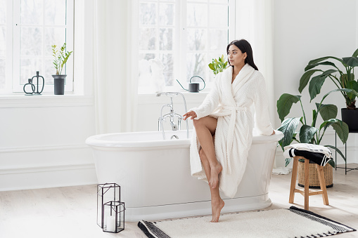 Body care, skincare and natural beauty concept. Full length view of young asian woman sitting on freestanding bath in white bathrobe, looking a side. Beautiful female in modern interior bathroom