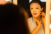 若いアジアの女性は、彼女の顔に顔のシートマスクを適用し、バスルームで彼女の反射を見ています