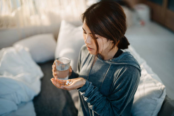 Young Asian woman sitting on bed and feeling sick, taking medicines in hand with a glass of water Young Asian woman sitting on bed and feeling sick, taking medicines in hand with a glass of water diabetes symptoms stock pictures, royalty-free photos & images