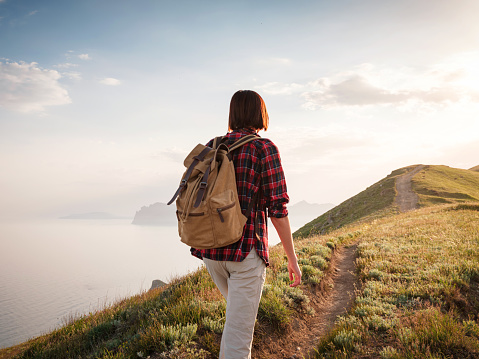 A young Asian woman with a backpack hiking in the summer climbing the mountain at sunset . Mountain and coastal travel, freedom and an active lifestyle
